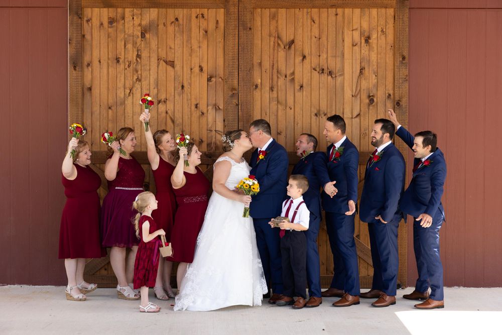 Wedding party shows playful expressions in burgundy and navy attire against rustic barn setting.