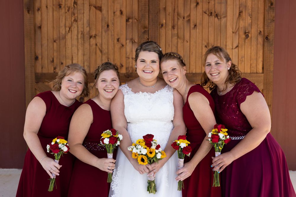 Bride and bridesmaids in burgundy dresses hold colorful sunflower bouquets against wooden backdrop.