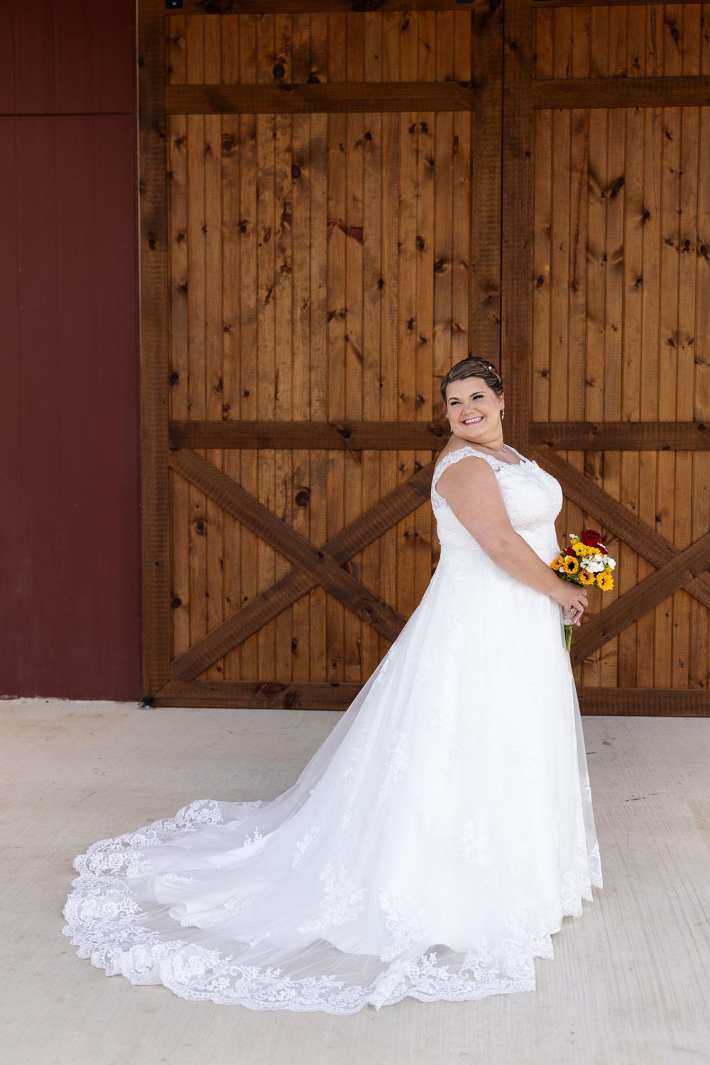 Bride in lace wedding gown with train poses against rustic wooden barn doors.