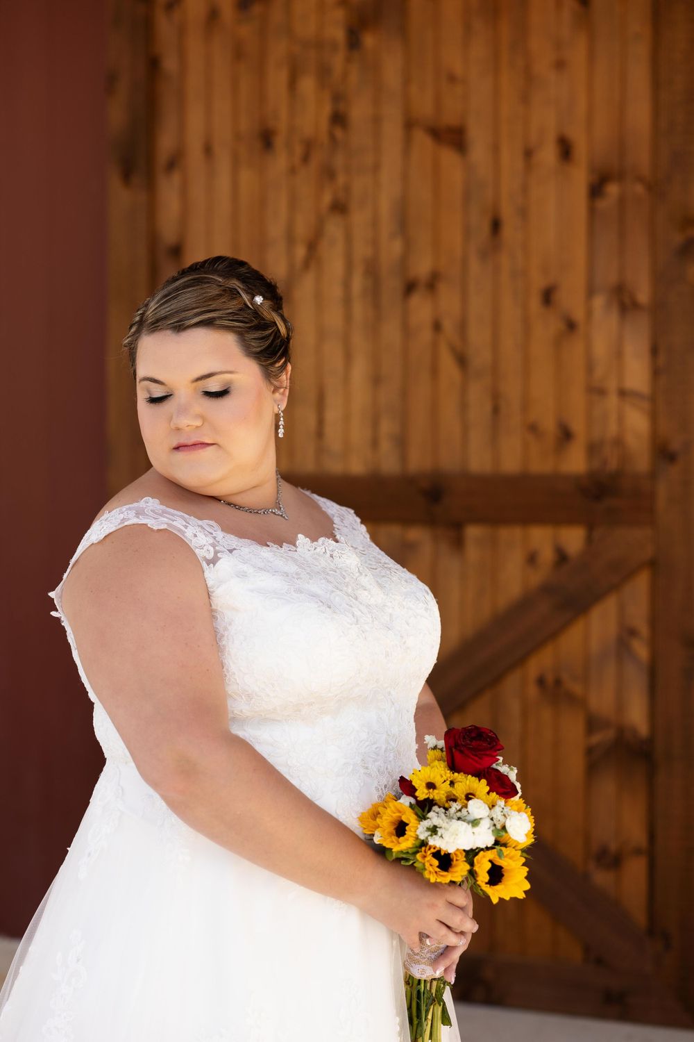 A bridal portrait in a white lace dress holding a sunflower and rose bouquet against a rustic wooden barn backdrop.