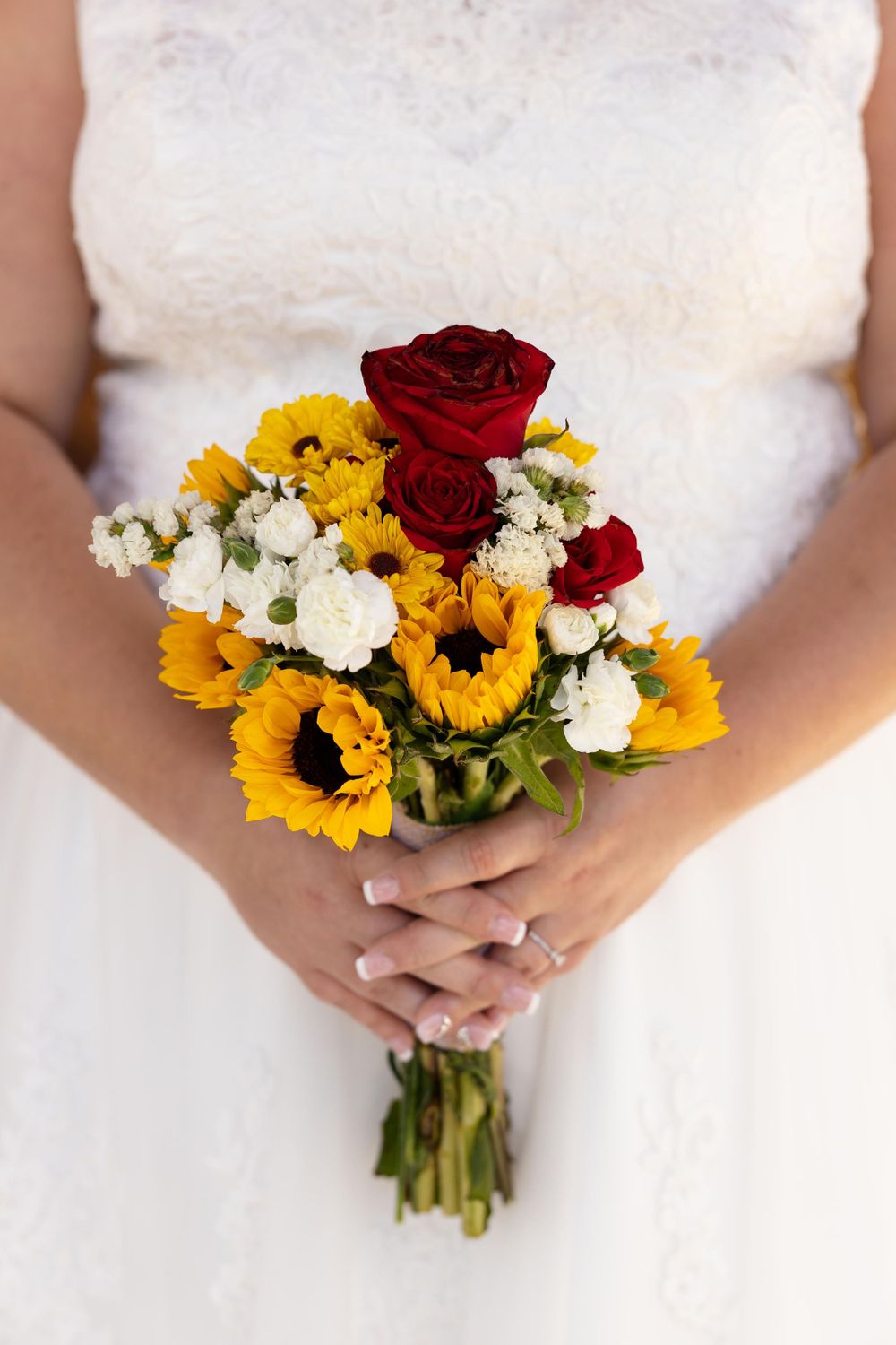 A vibrant bridal bouquet featuring sunflowers, red roses, and white flowers against a white wedding dress.