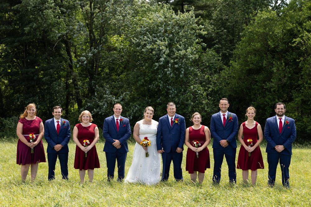 Wedding party in burgundy dresses and navy suits lined up outdoors against green foliage.