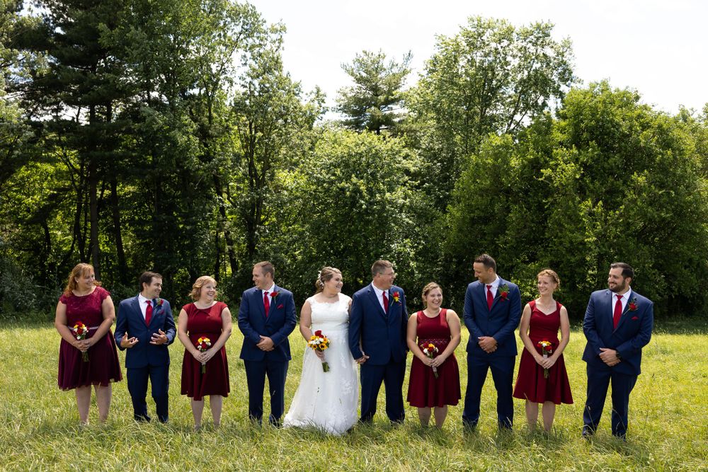 Wedding party stands in a row on grassy field with bridesmaids in burgundy and groomsmen in navy suits.