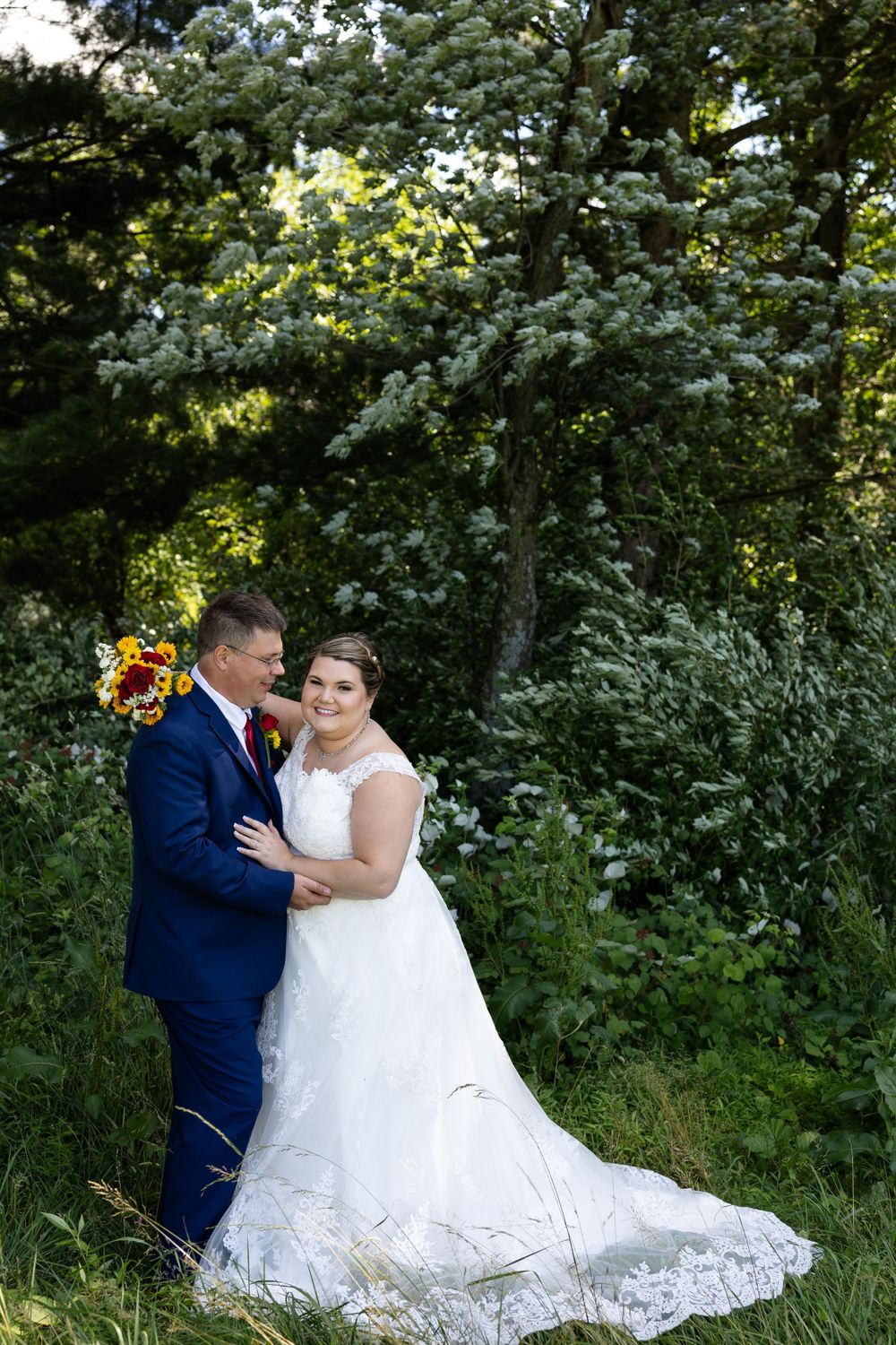Wedding couple poses against lush greenery with sunflower bouquet and flowing lace train.