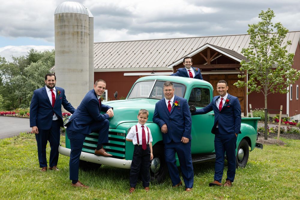 A group stands beside a vintage mint green Chevrolet pickup truck on a farm with a red barn and silo in the background.