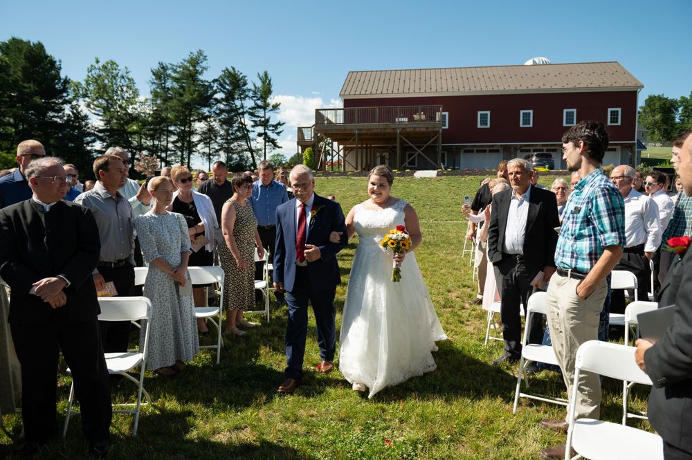 Wedding guests gather on a lawn near a rustic brown barn during an outdoor ceremony.