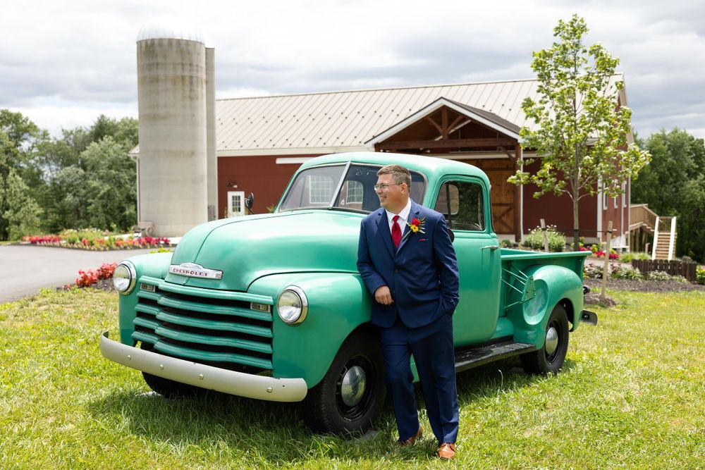 A classic mint green Chevrolet pickup truck parked in front of a red barn with silo on a cloudy day.