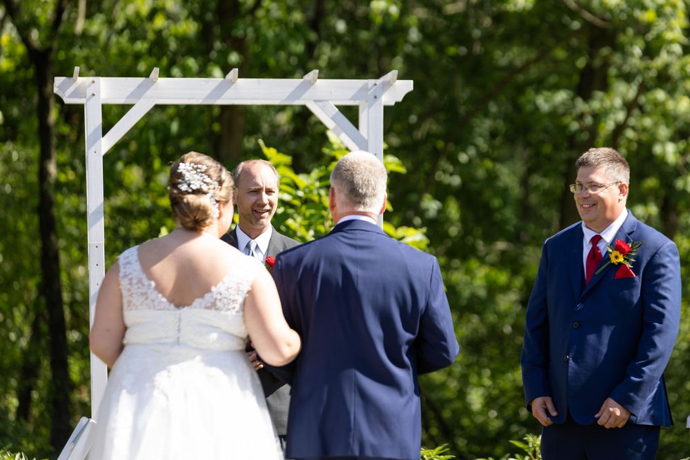 An outdoor wedding ceremony under a white wooden arch surrounded by lush greenery on a sunny day.