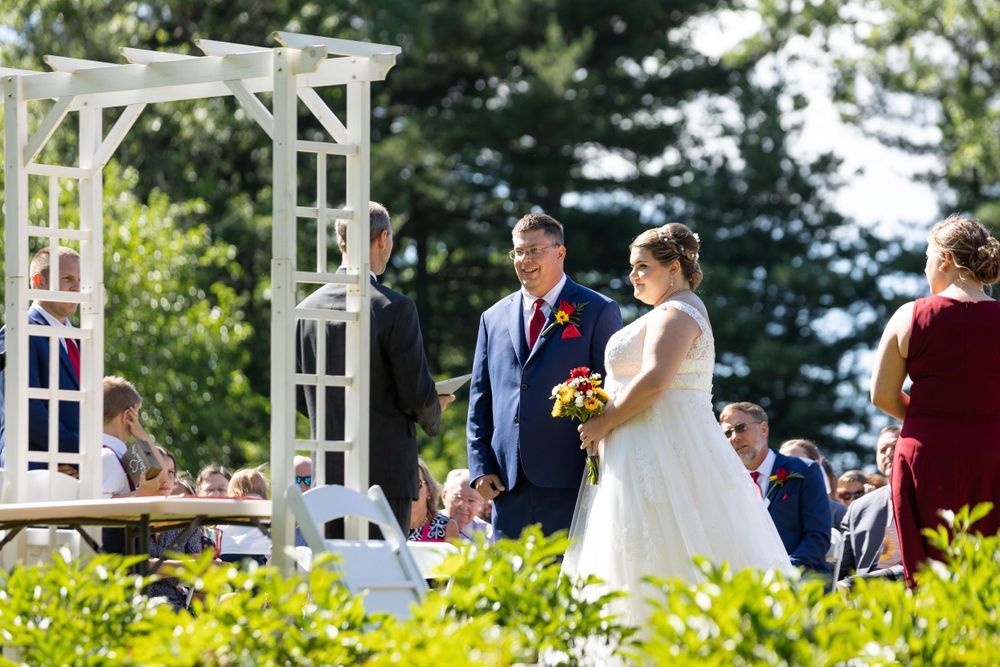 Wedding ceremony taking place under a white pergola with bright sunlight and evergreen trees in background.
