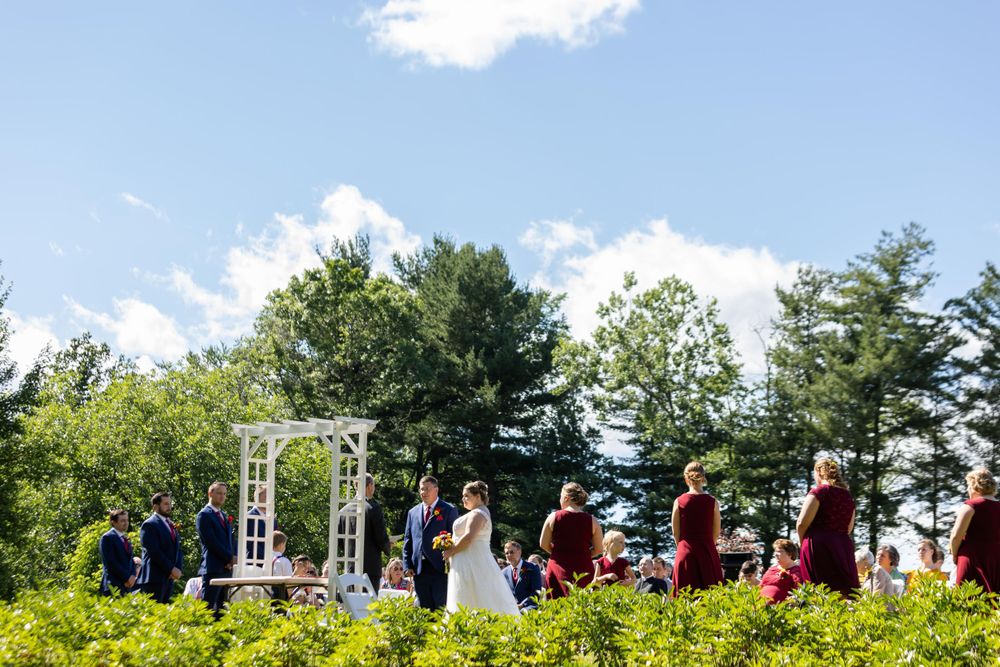 Wedding ceremony viewed from afar showing white arch and gathered guests against blue summer sky.