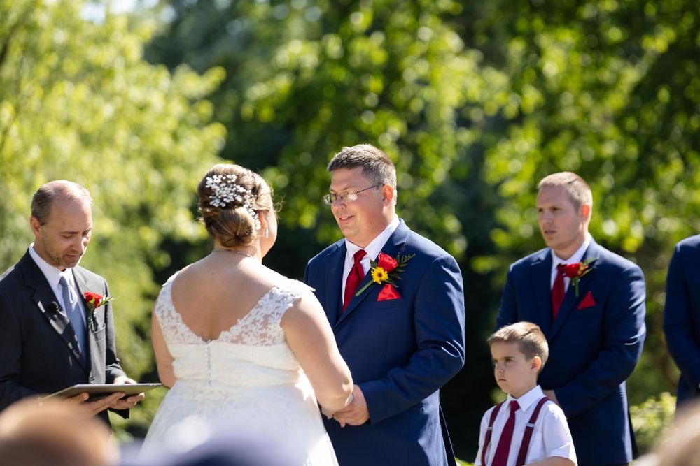 Outdoor summer wedding ceremony with attendants in navy suits and red boutonnieres.