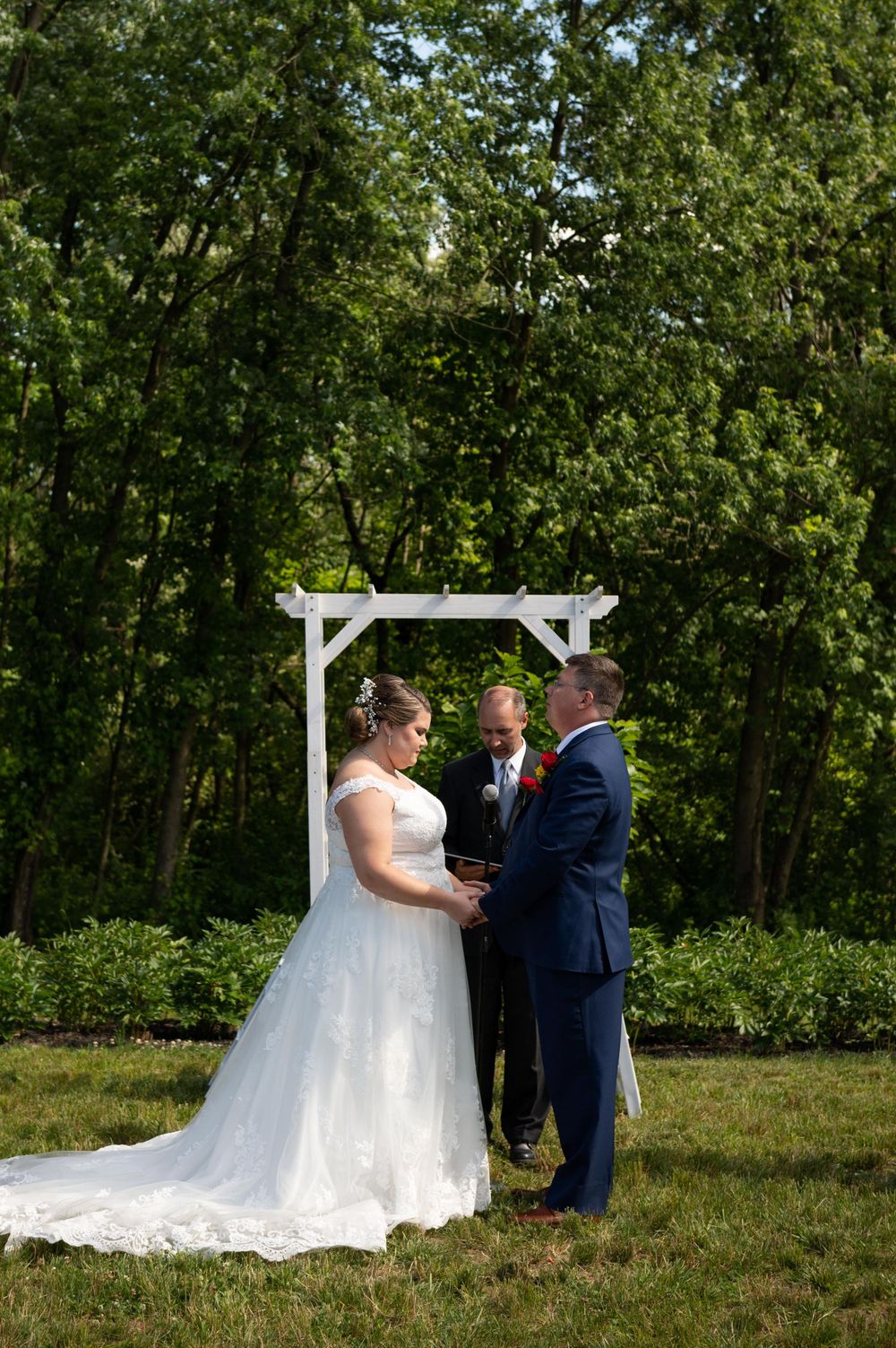 Wedding ceremony under white wooden arch with dense forest backdrop on sunny afternoon.
