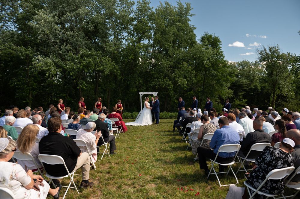Wide view of outdoor wedding ceremony with white chairs lined up between trees on grassy field.