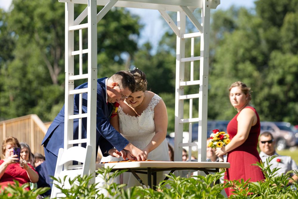 Wedding couple signs marriage documents under white lattice arch during outdoor ceremony.