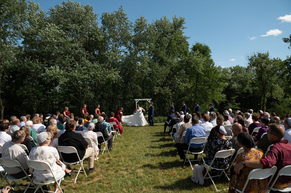 A sunny outdoor wedding ceremony with guests seated in white chairs on a grassy lawn surrounded by trees.