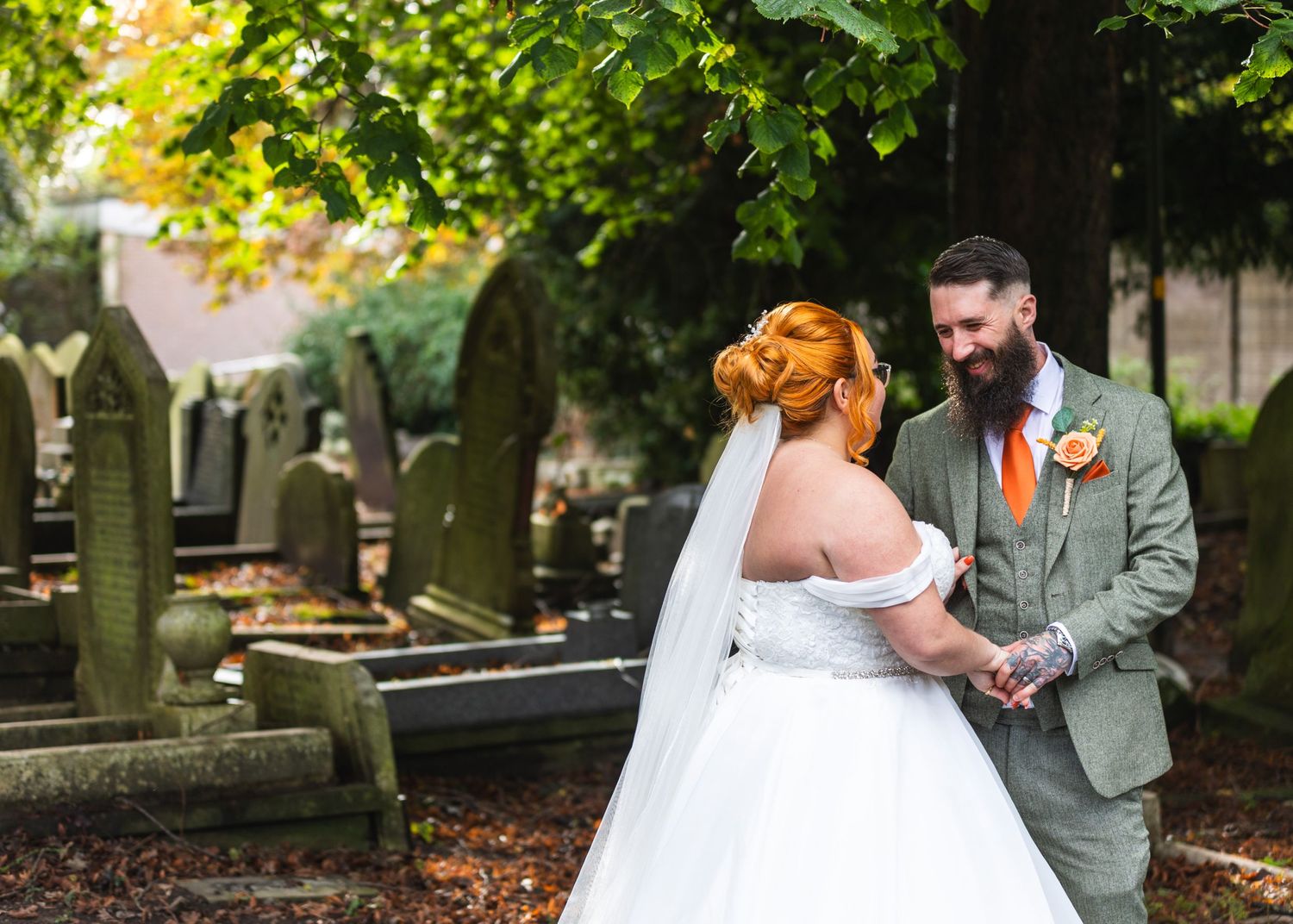 A bride in a white dress and groom in a gray suit share an intimate moment in a historic cemetery setting.