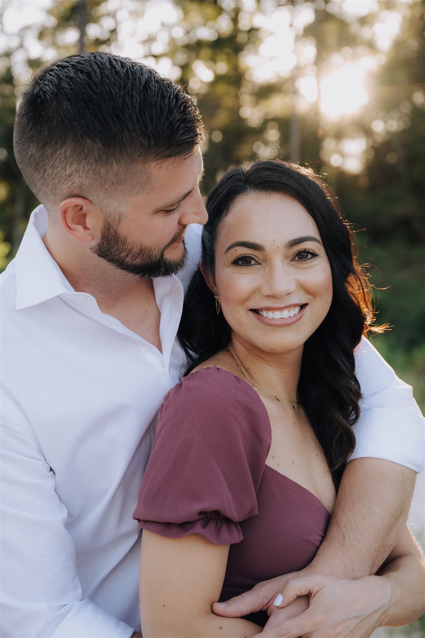 Close up portrait of couple in burgundy dress and white shirt smiling together in golden sunlight.