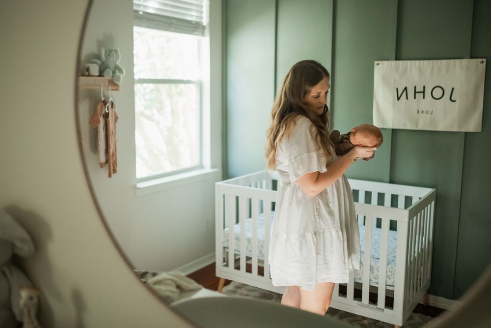 Woman in white dress stands in mint green nursery room looking at white crib and baby items.