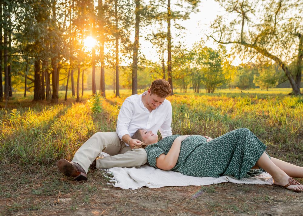 A couple in white and green outfits share a moment on a blanket in a sunlit forest with golden wildflowers.