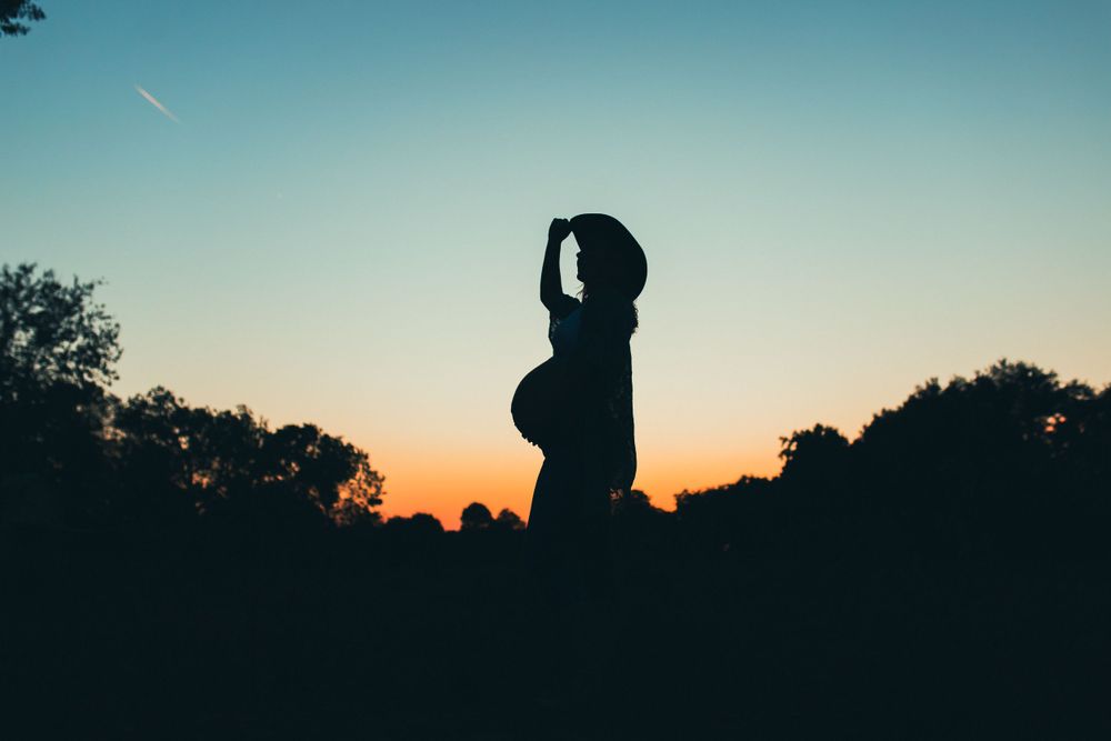 A pregnant silhouetted figure stands against a vibrant orange and blue sunset sky with trees in the background.