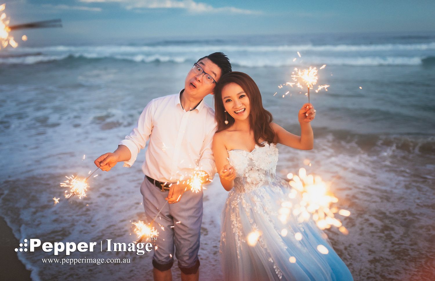 Couple holding sparklers on beach at sunset in wedding attire.