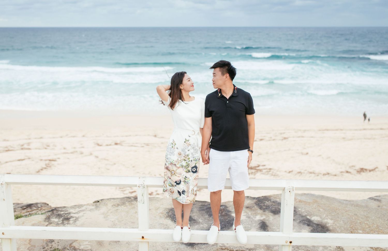 Couple in casual attire hold hands and smile at each other on a sunny beachside fence.