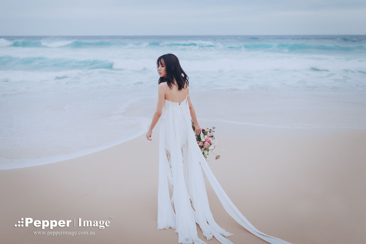Bride in elegant white gown holding bouquet on sandy beach with ocean waves.