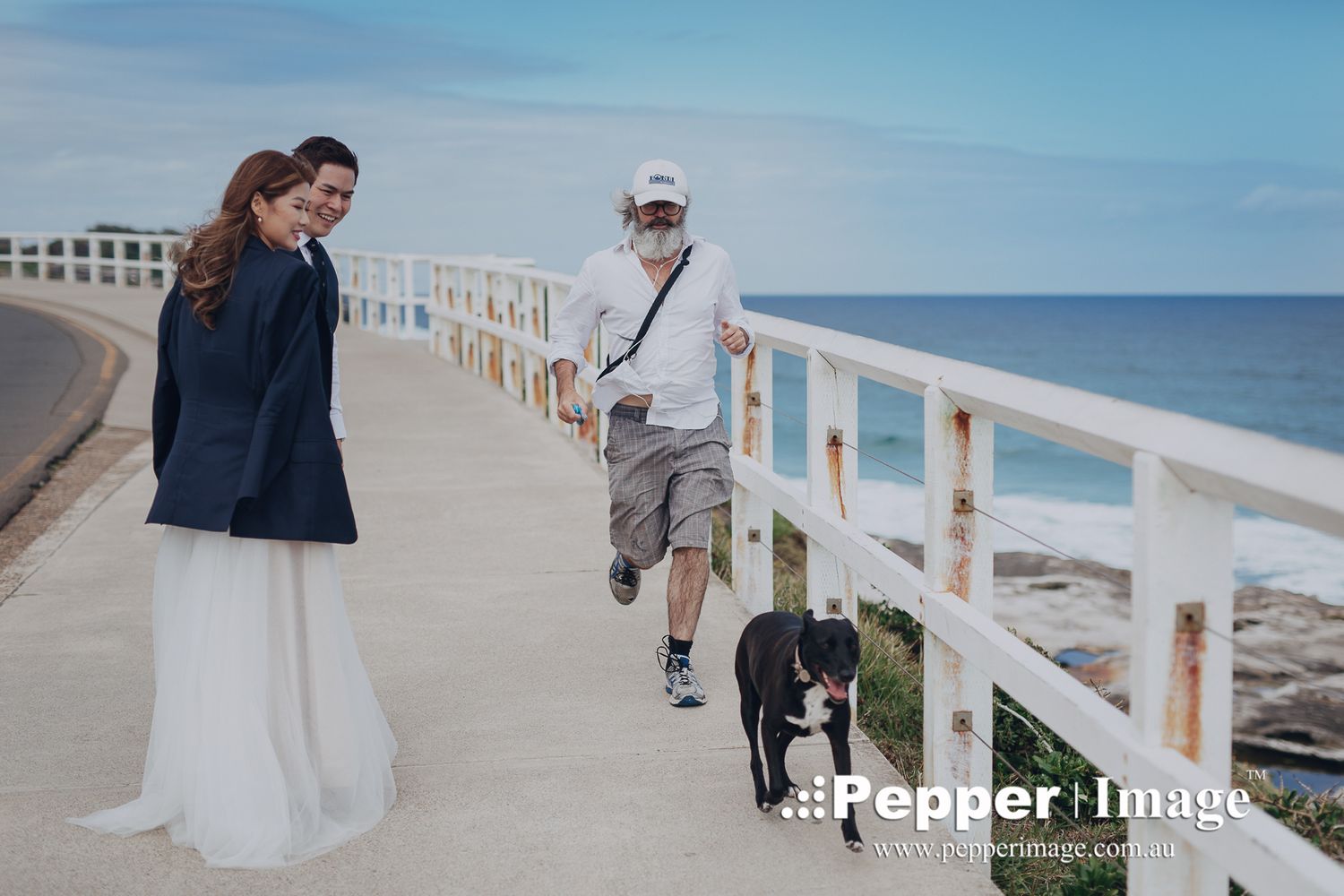 Bride and groom on coastal walkway as jogger passes with black dog on sunny day.