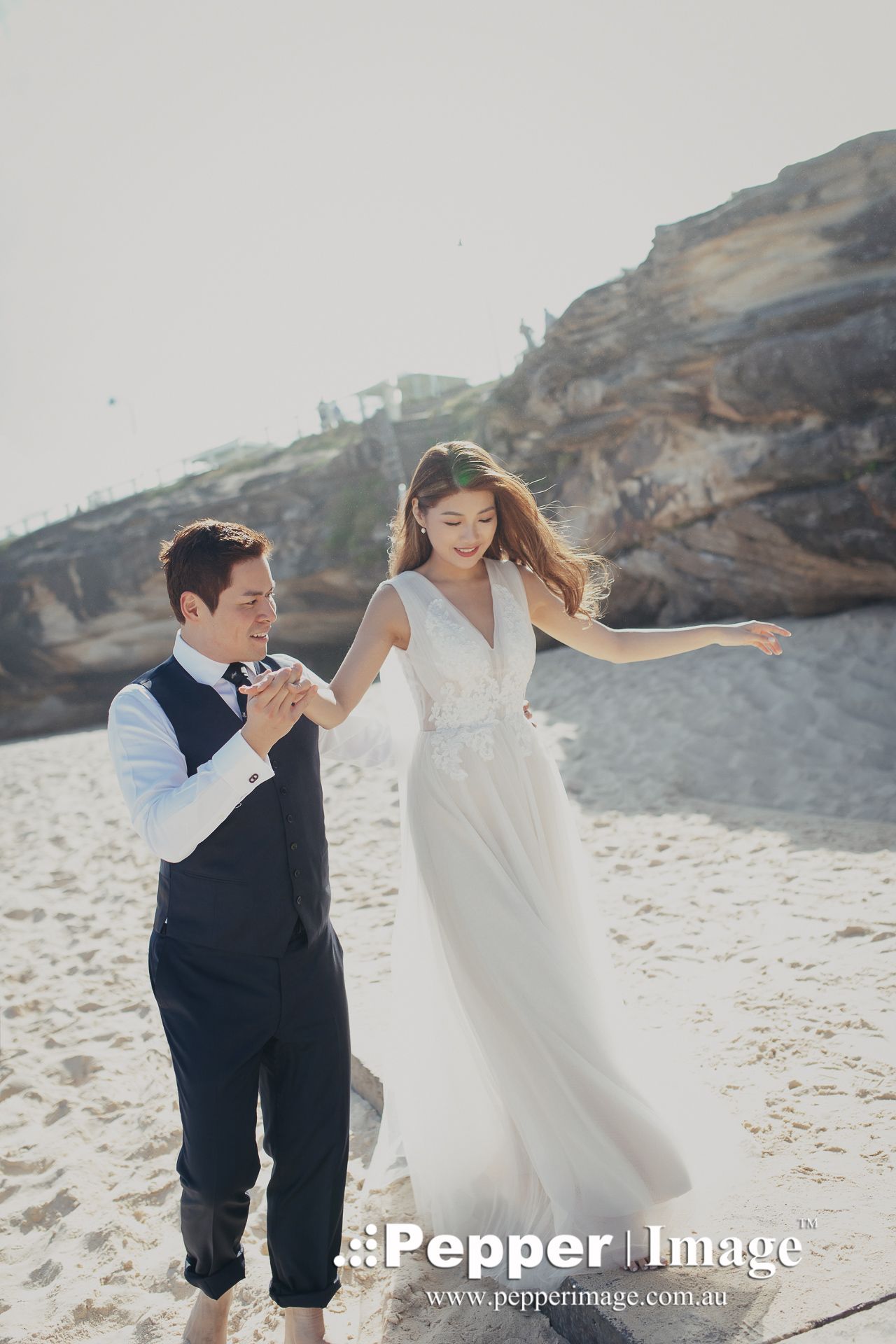 Groom twirls bride in flowing white dress on sandy beach with rocky cliffs behind them.