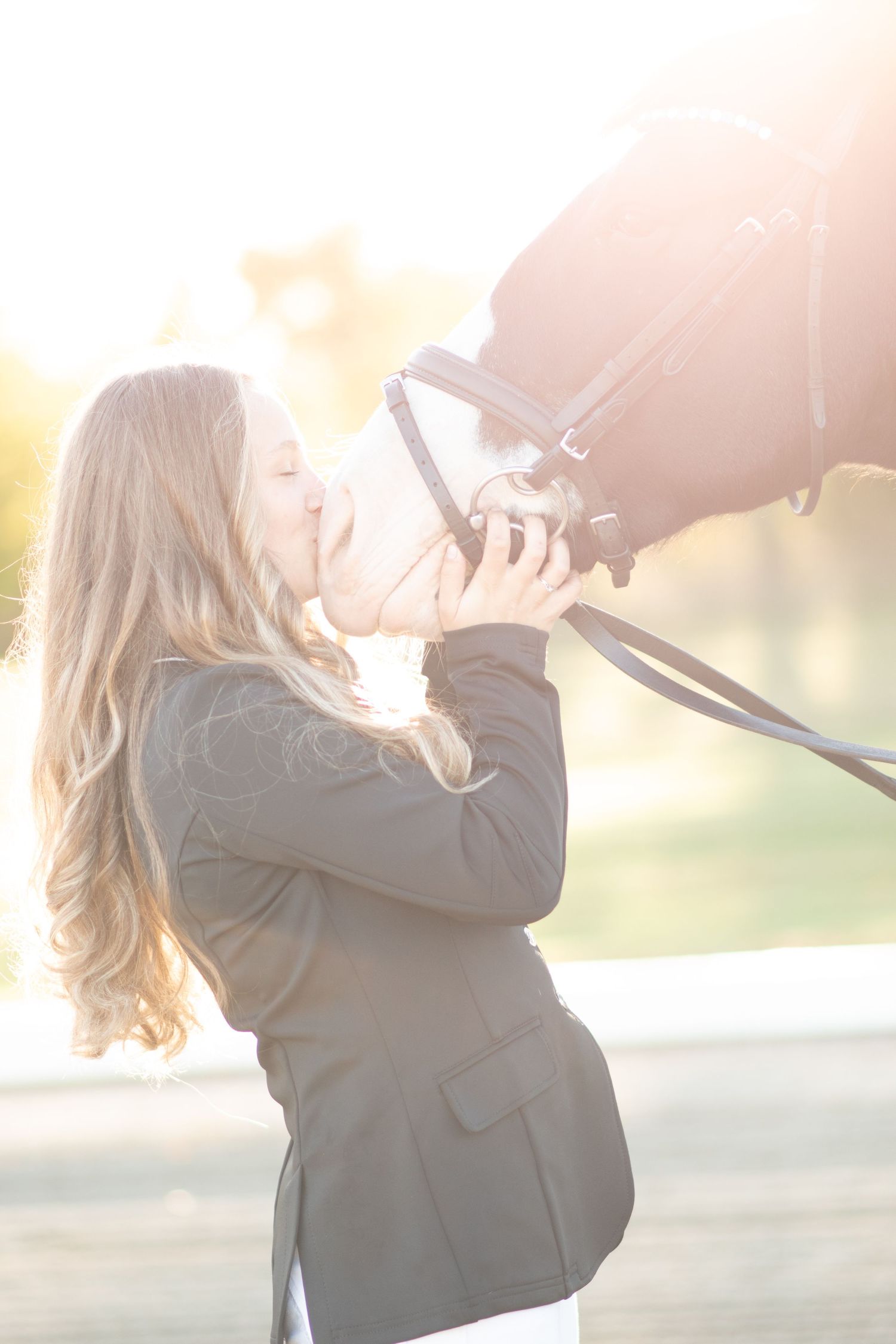 Young blonde woman kissing a dark horse on the nose in soft golden backlit outdoor light. Equestrian Photographer in Maine.