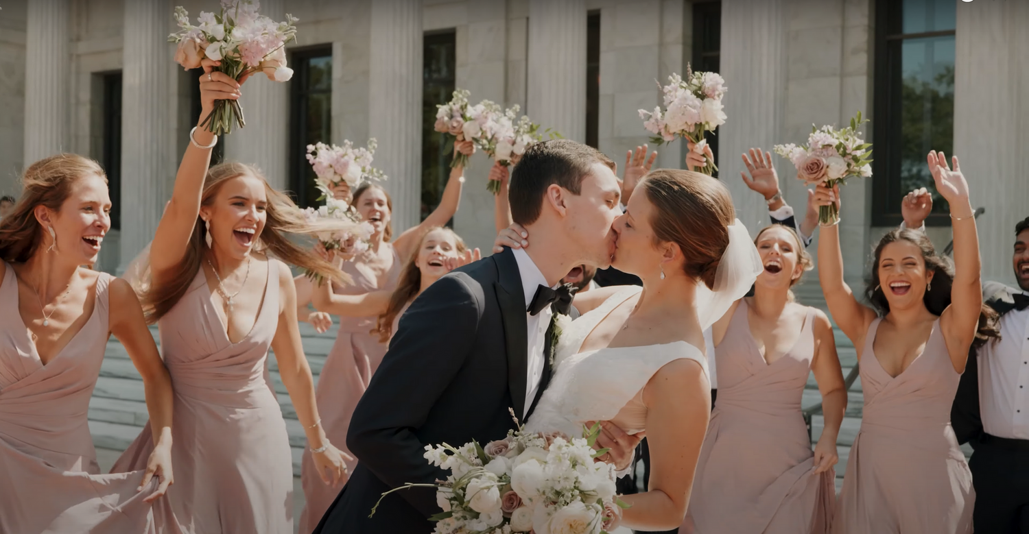 Wedding party celebrates with bouquets raised as the newlyweds share a kiss.