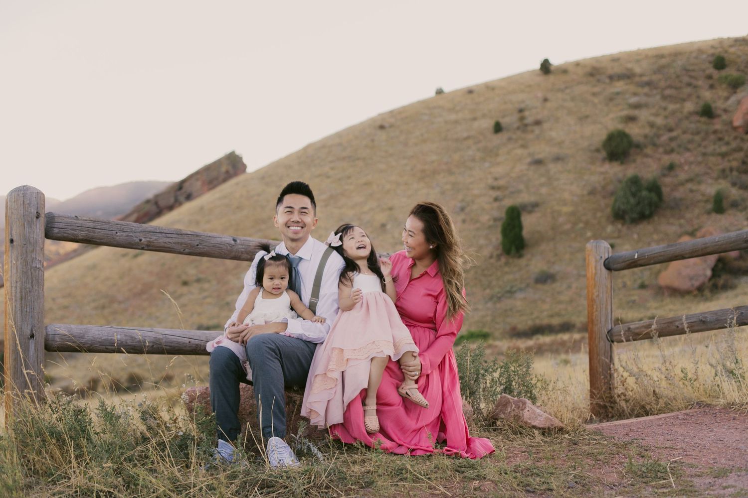Family sitting together on wooden fence in casual attire against rolling hills at sunset.
