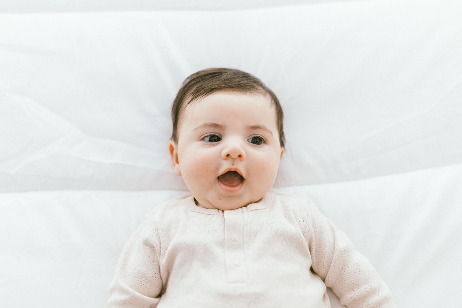 Baby in pale pink outfit lying on white sheets with mouth open in excitement.