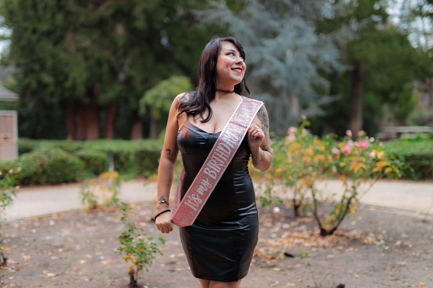 Woman in black dress wearing graduation sash poses in garden setting.
