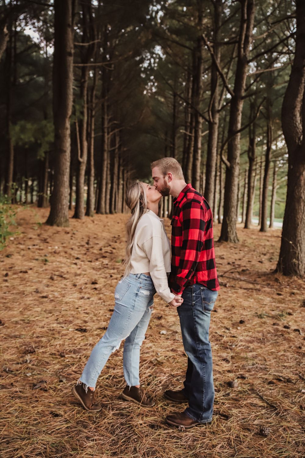 A romantic couple shares an intimate moment among tall pine trees in a forest setting.