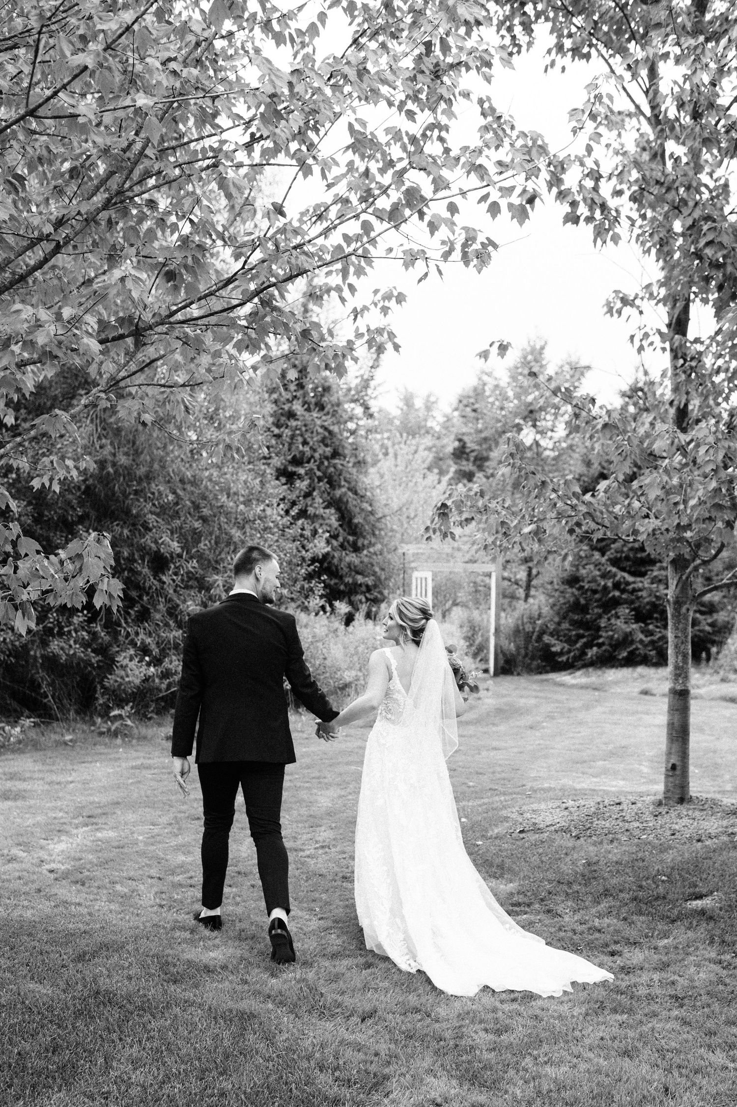 A couple walks hand in hand through a tree-lined path in black and white.