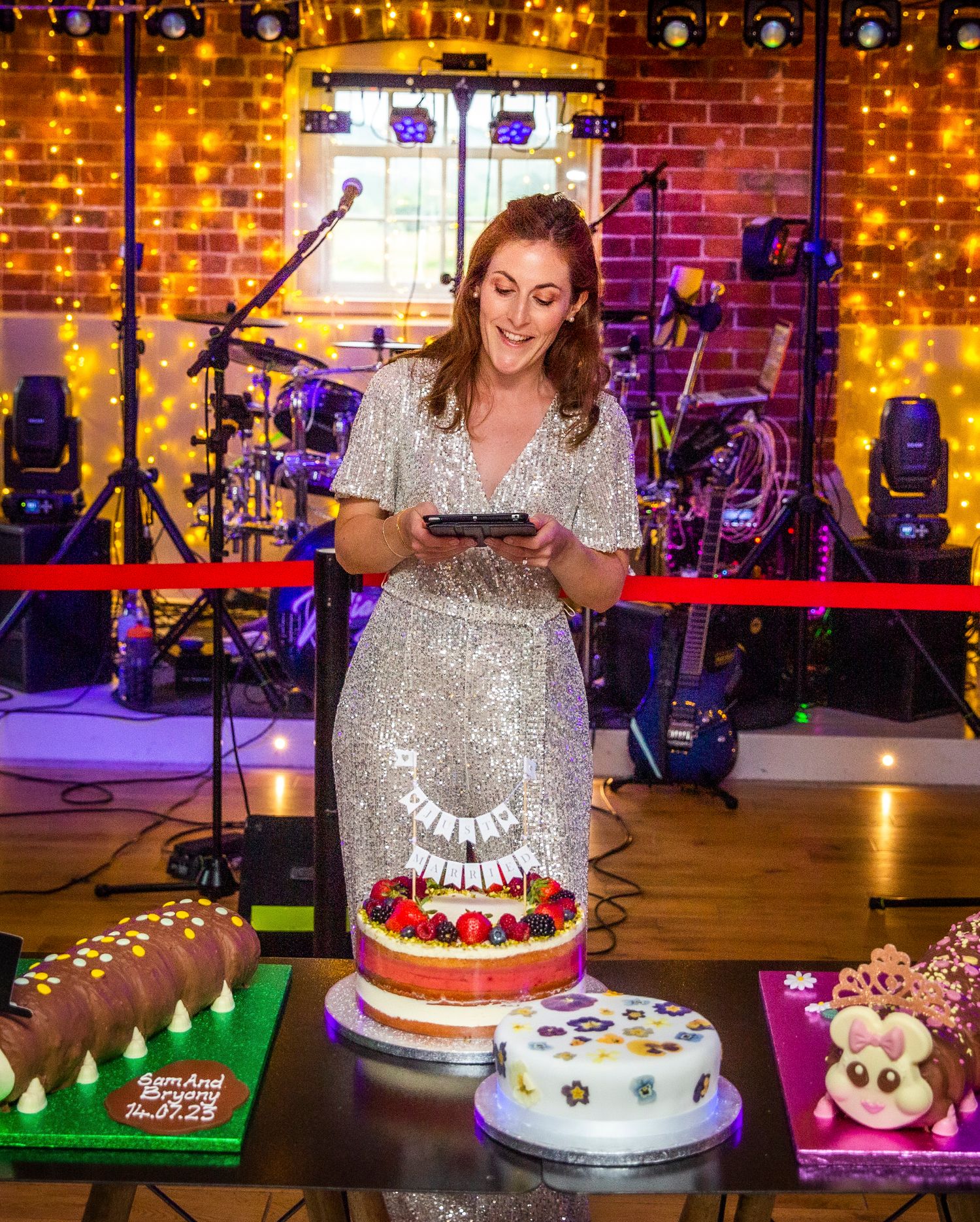 A sparkly silver dress stands out against a backdrop of string lights and musical equipment while cutting a festive cake.