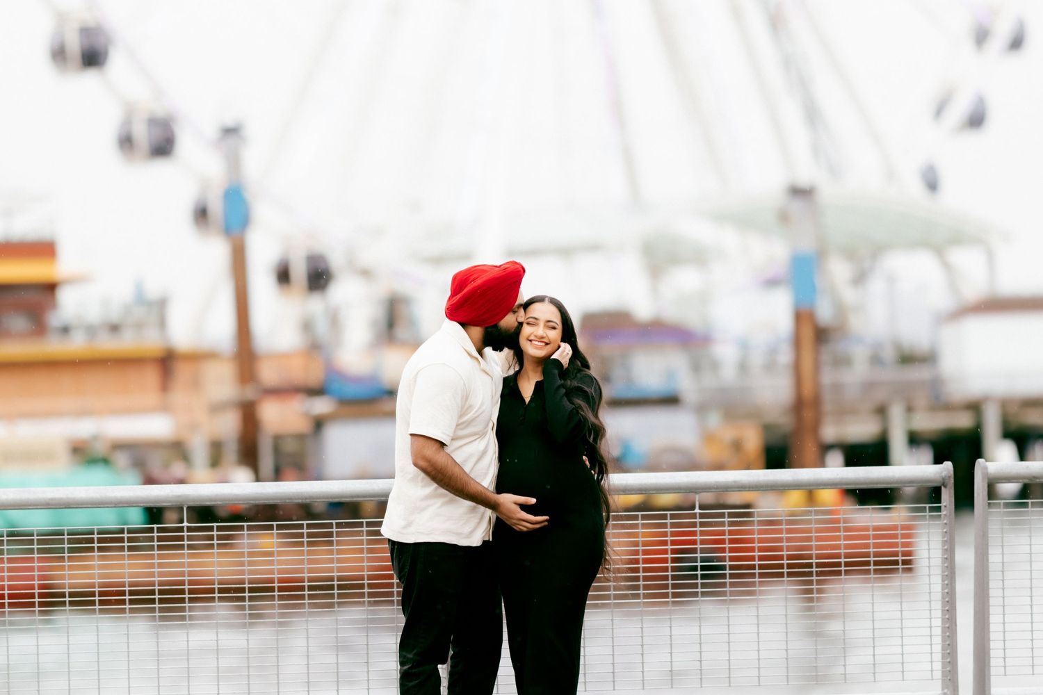 A couple embraces on a pier with amusement park rides visible in the background.