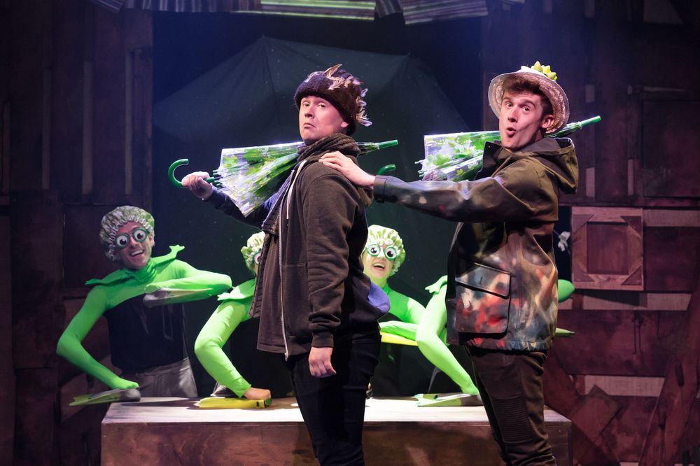 Two actors perform with bright green puppet props on a dark theatrical stage.
