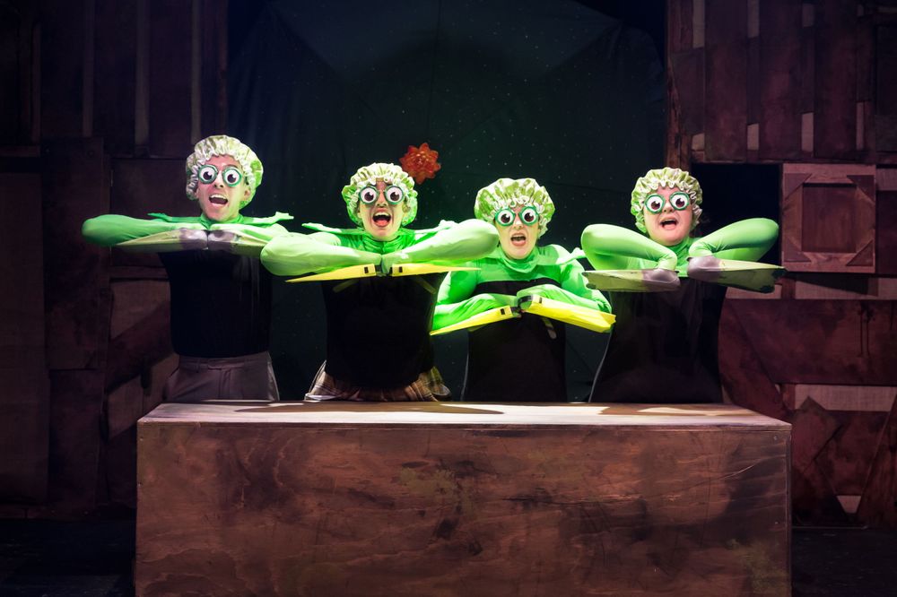Four performers with green props create a synchronized theatrical scene on stage.