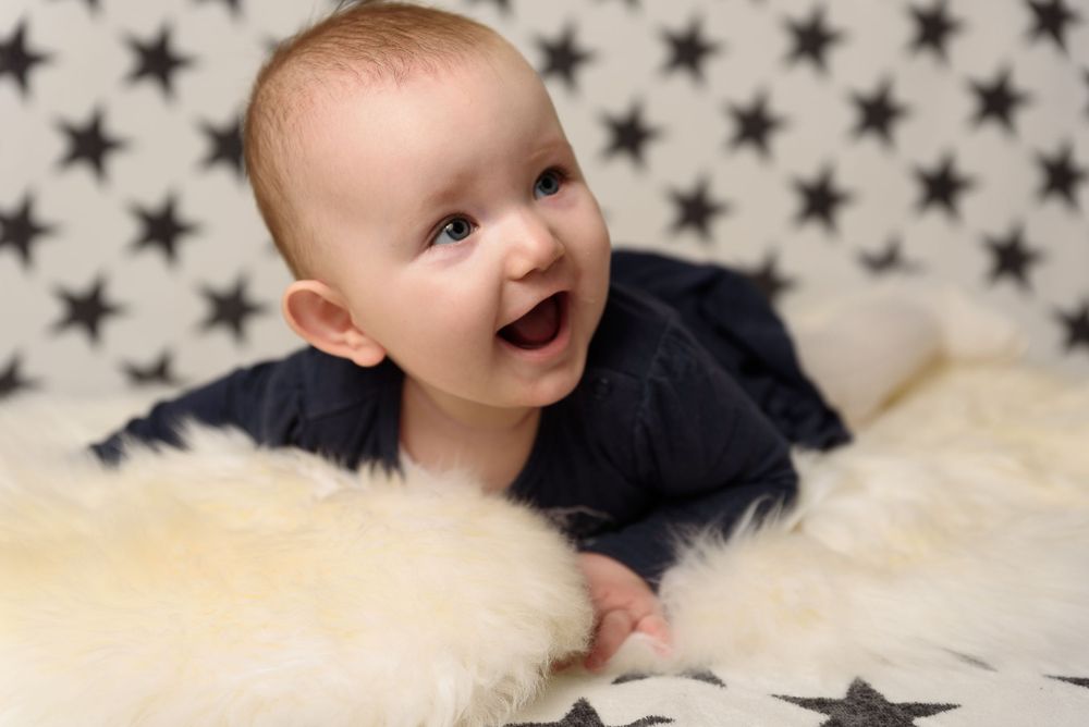 Smiling baby in dark outfit doing tummy time on fluffy white rug with star patterned background.
