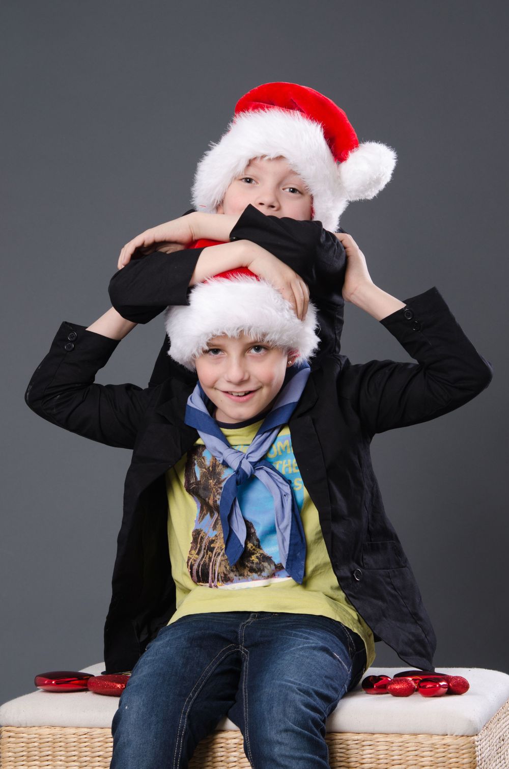 Two children wearing Santa hats posing playfully together for a Christmas portrait on grey background.