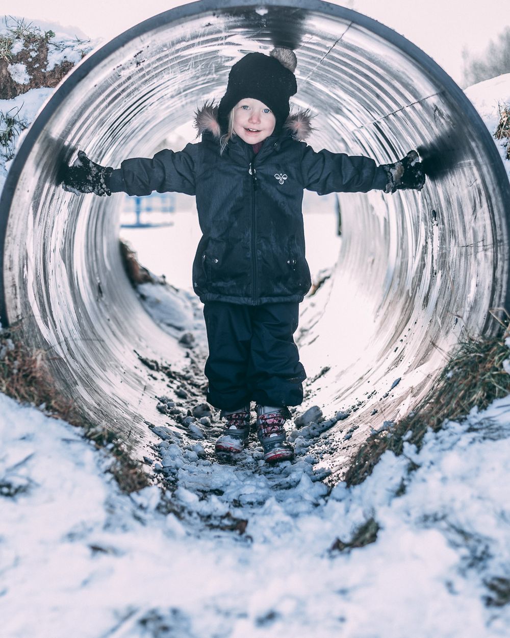 Happy toddler in black snowsuit standing inside a metal tunnel playground structure in winter snow.