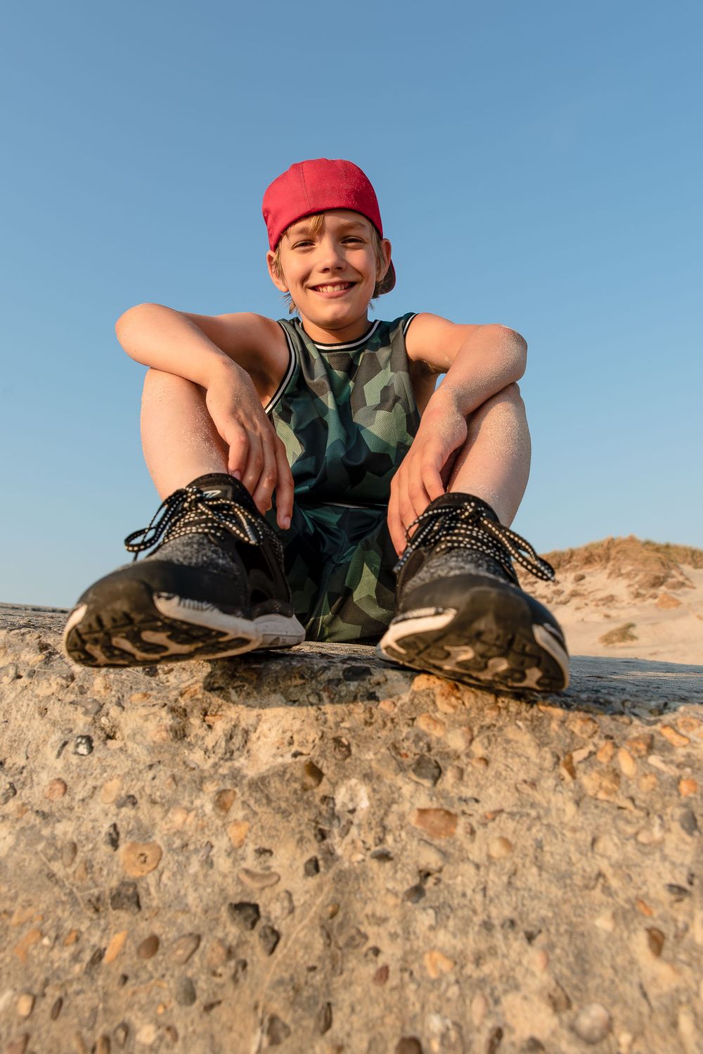 Smiling boy in red bandana and camo outfit sitting relaxed on a sandy rock under blue sky.