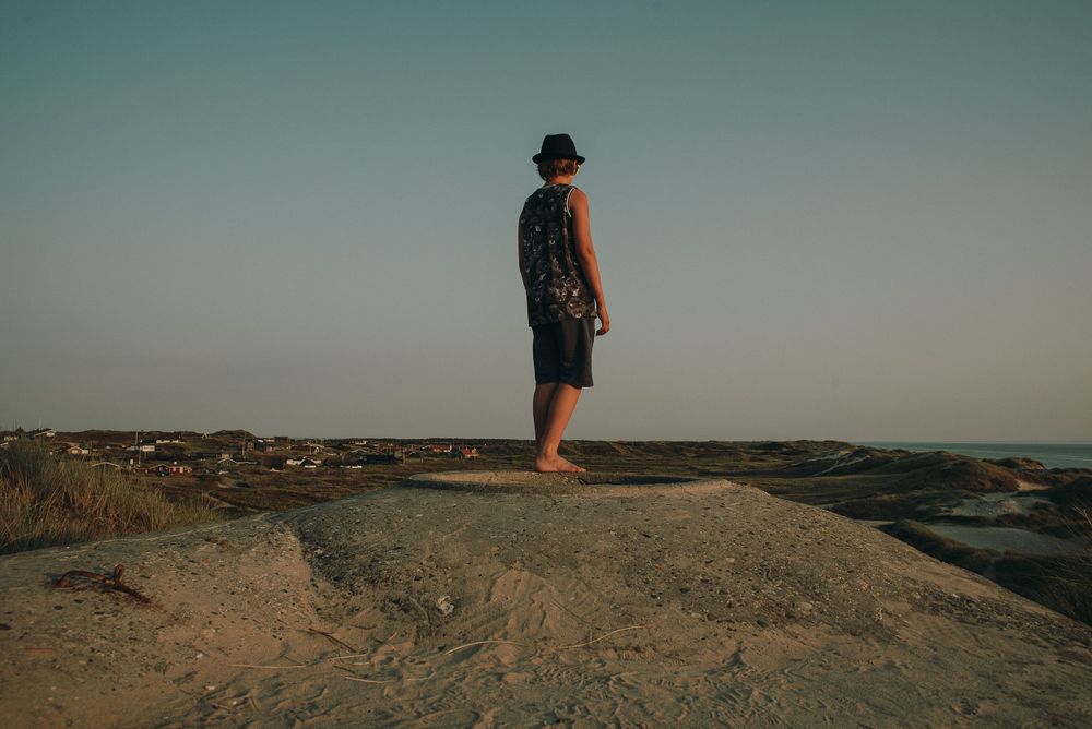 Lone man in a hat standing barefoot on a rocky coastal outcrop gazing at the horizon at dusk.