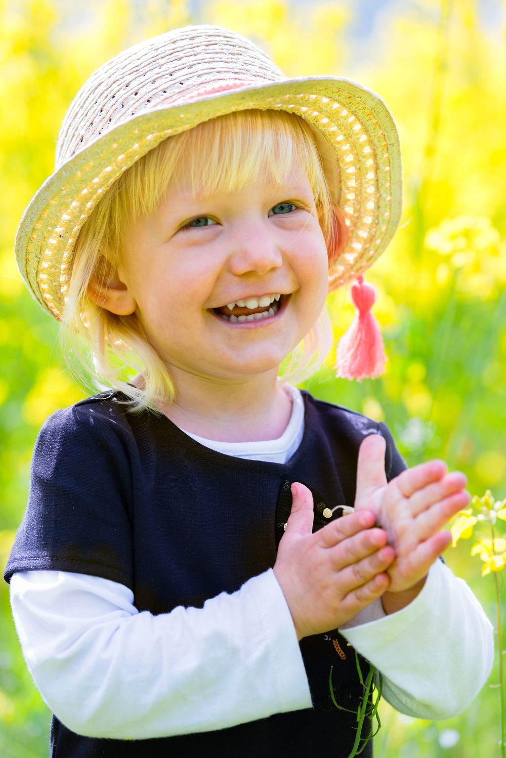 Happy blonde toddler in a straw hat clapping hands in a bright yellow flower field.