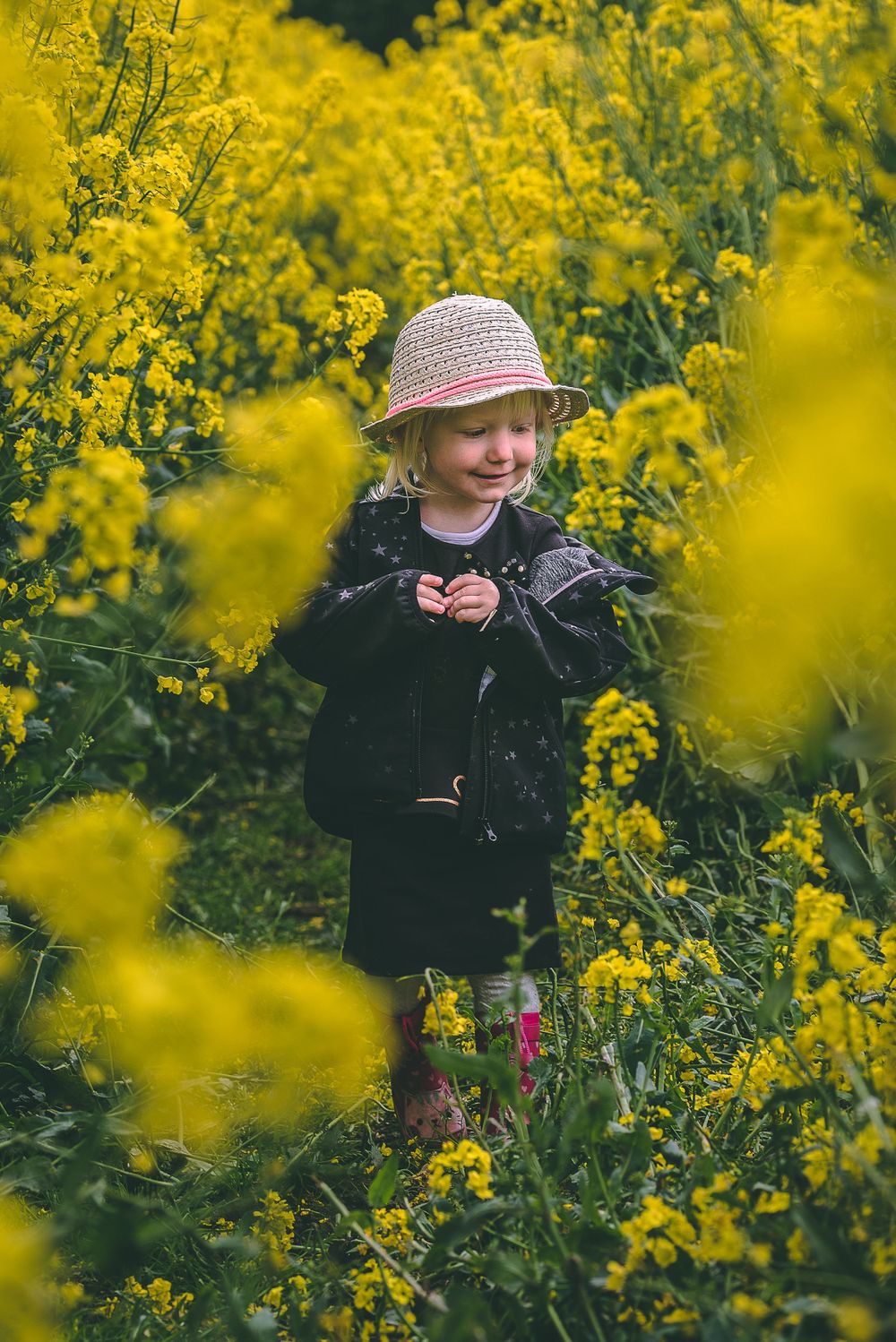 Young girl in straw hat and black jacket smiling among bright yellow rapeseed flowers in a field.