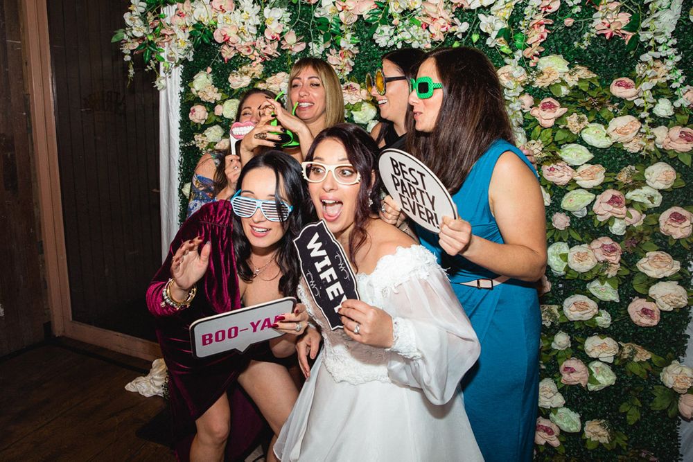 Wedding party members posing with props against a floral backdrop during reception celebrations.