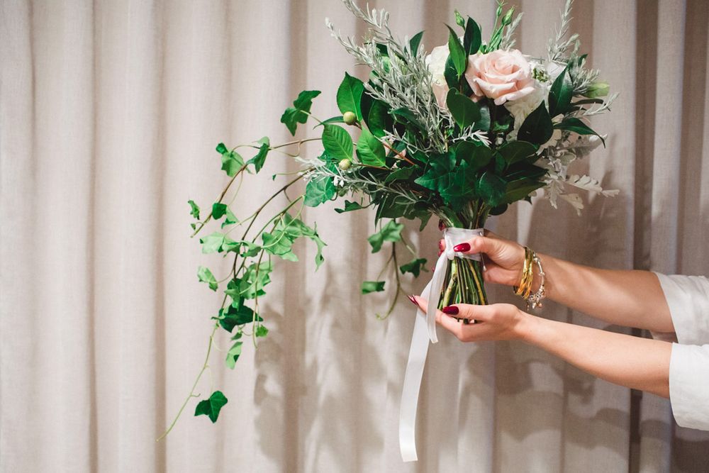 Wedding bouquet with pink roses and greenery held against a light curtain backdrop.