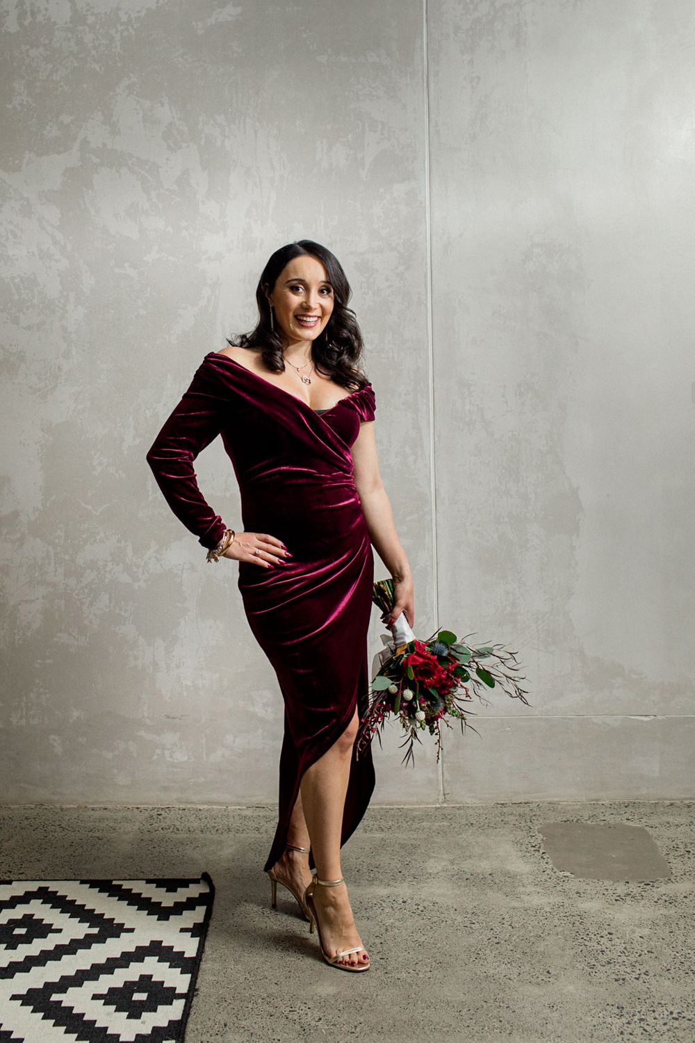 Fashion poses in burgundy velvet dress against industrial grey backdrop.
